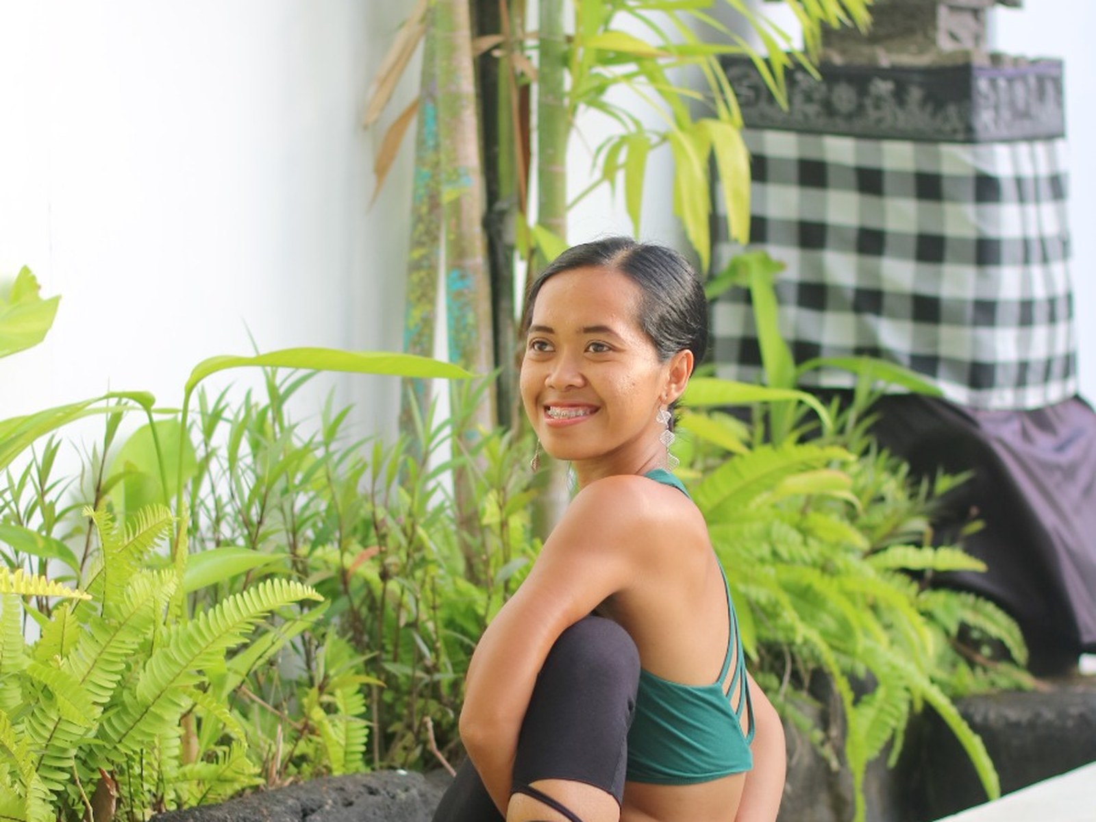 Smiling woman in a seated yoga pose beside a pool, among tropical plants in Bali
